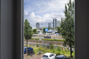 Uitzicht vanuit een raam op de De Ruijterkade in Amsterdam met geparkeerde auto's, treinspoor, bomen en moderne hoogbouw op de achtergrond.